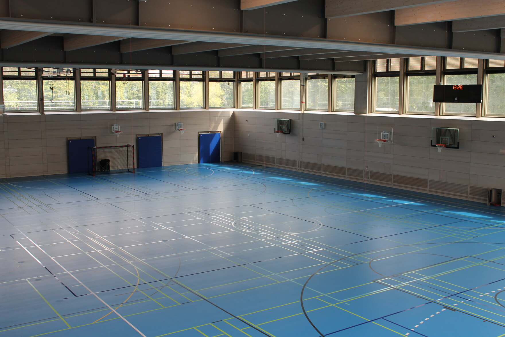 Die Wände im Inneren der Turnhalle sind in hellem Holz gehalten und der Boden besteht aus blauem Belag. Eine umläufige Fensterreihe im oberen Bereich lässt viel Licht ins Innere.